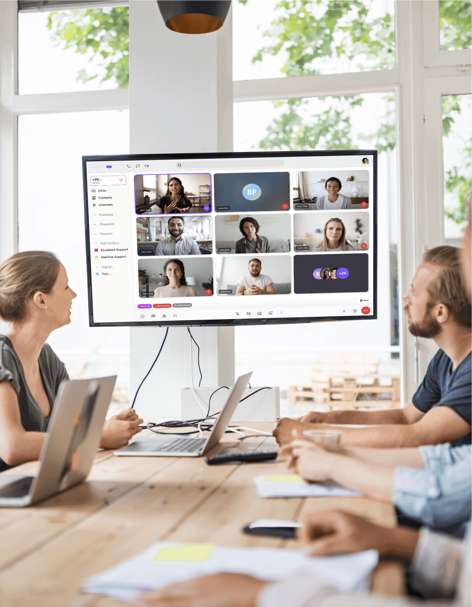 A large computer monitor in a conference room showing a video conference in progress with in-person and virtual attendees