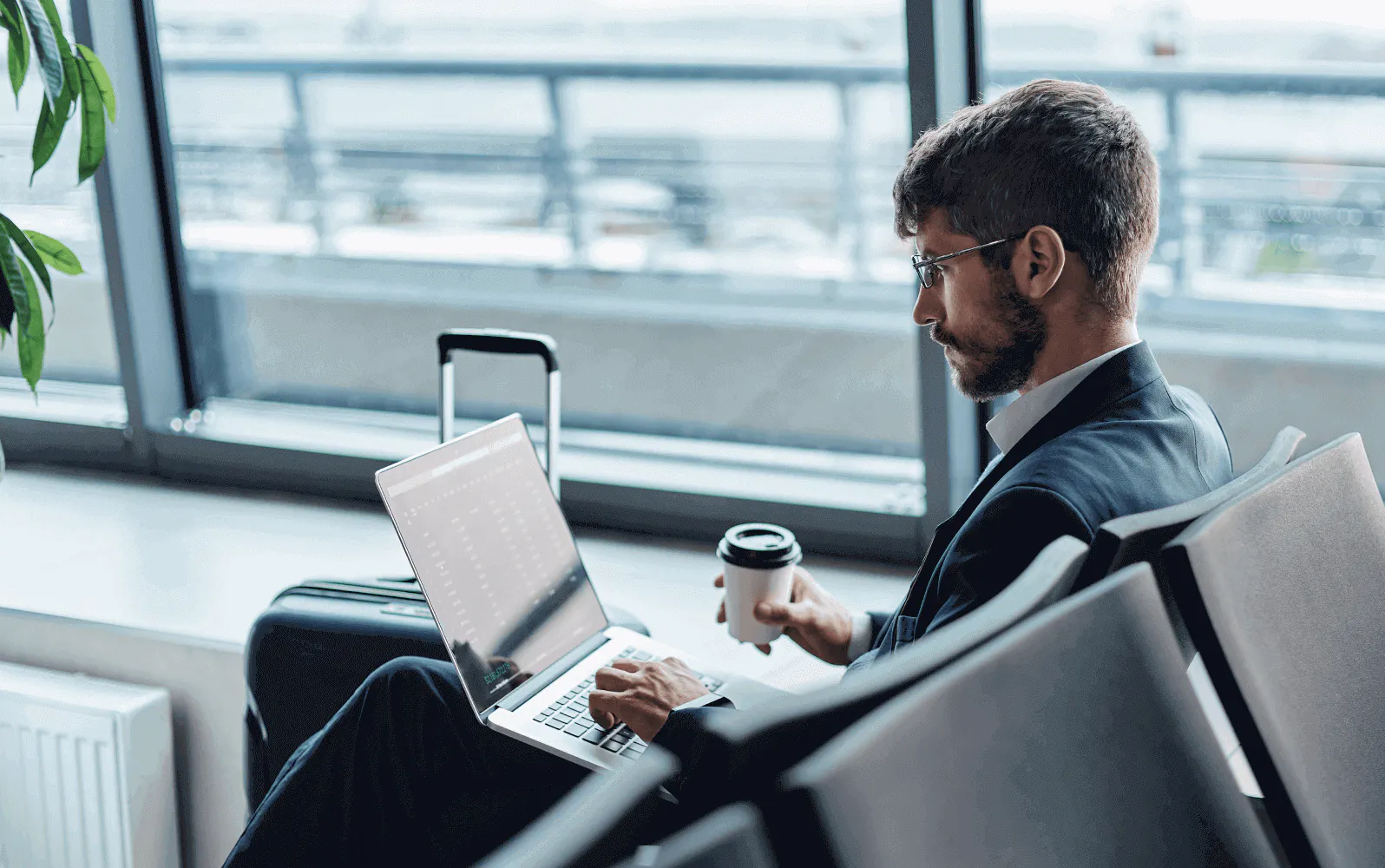 Man sitting in an airport waiting area with his laptop out and coffee in hand